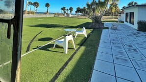 A view from inside a building looking out onto a grassy area with a few white chairs and a small table, a clear blue sky, and palm trees in the distance. - 3 Bedroom 3 Bath  a waterfront 3 Acre Home close to Downtown and Beaches. (Fort Pierce)