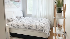 Ein Schlafzimmer mit Bett, einer Kandelaber, einem Fenster mit Vorhängen und einem an der Wand befestigten Regal mit Pflanze und Dekoration.