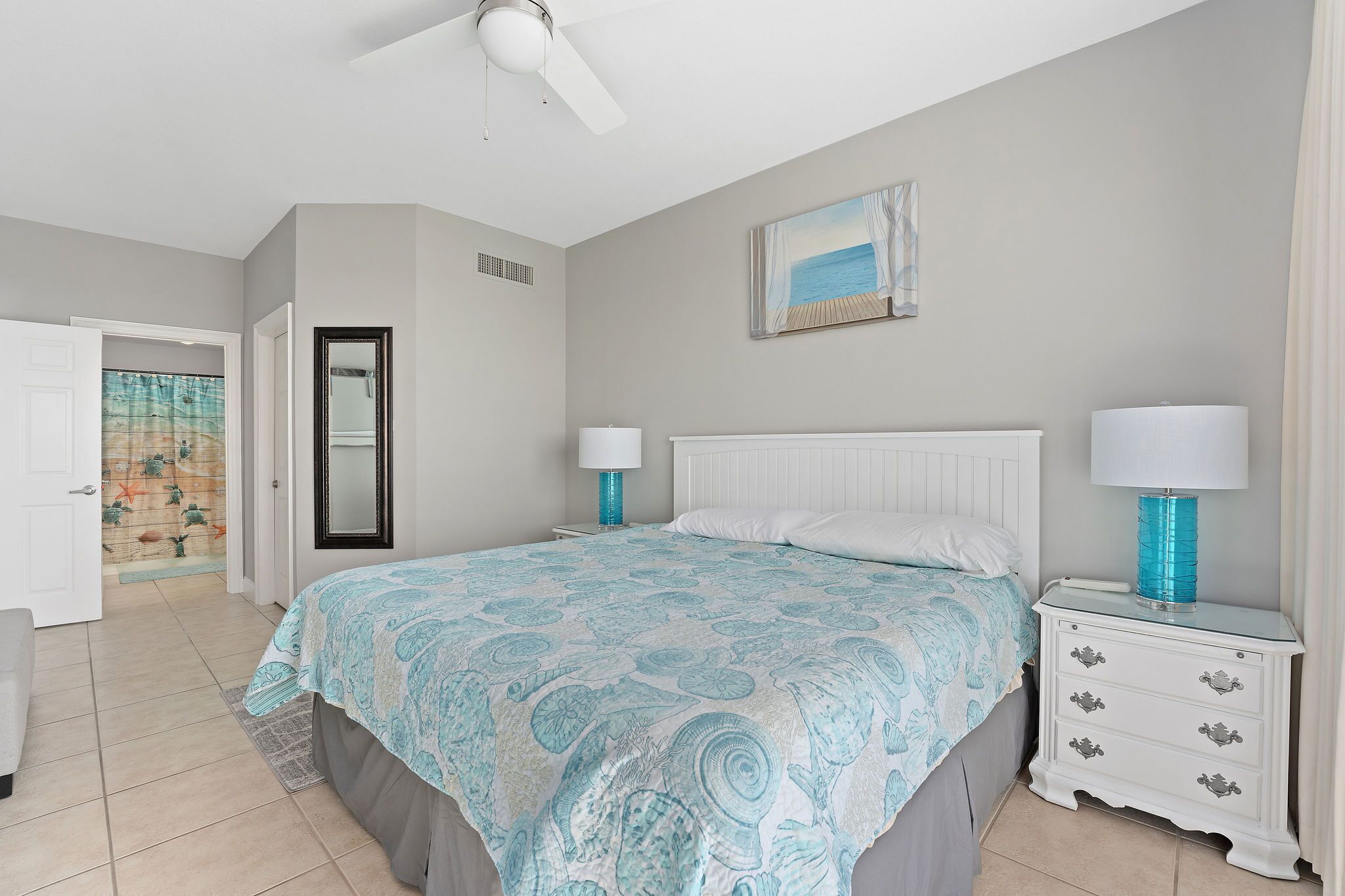 A bedroom with a bed, nightstands, a ceiling fan, a door leading to another room, and a painting on the wall.