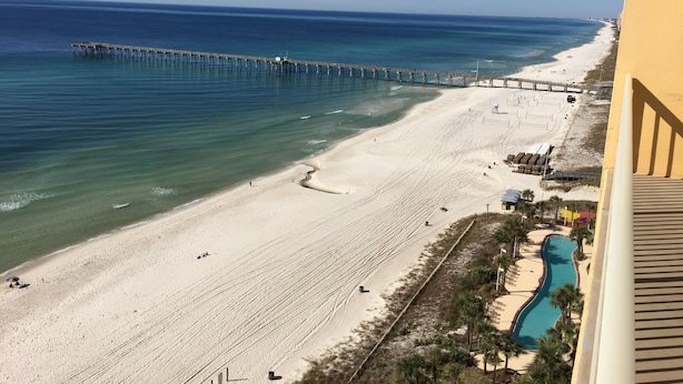 Una playa con un muelle que se adentra en el océano, arena y un área de piscina con sombrillas.