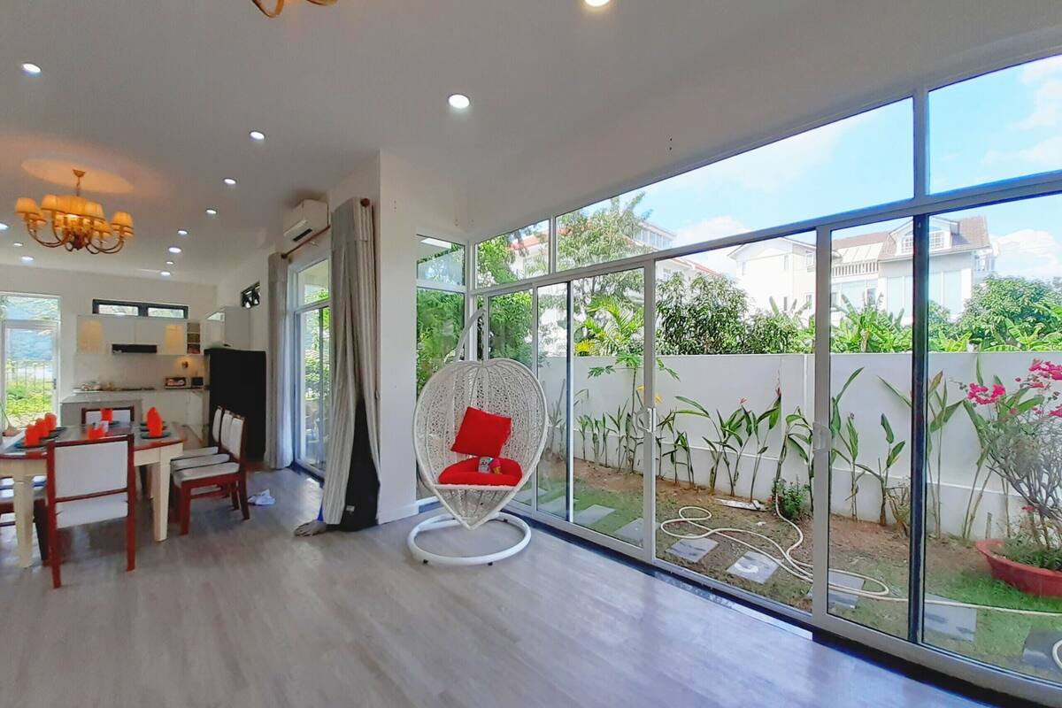 A spacious living area with a dining table, chairs, a hanging chair, and large windows offering a view of the garden and neighboring houses.