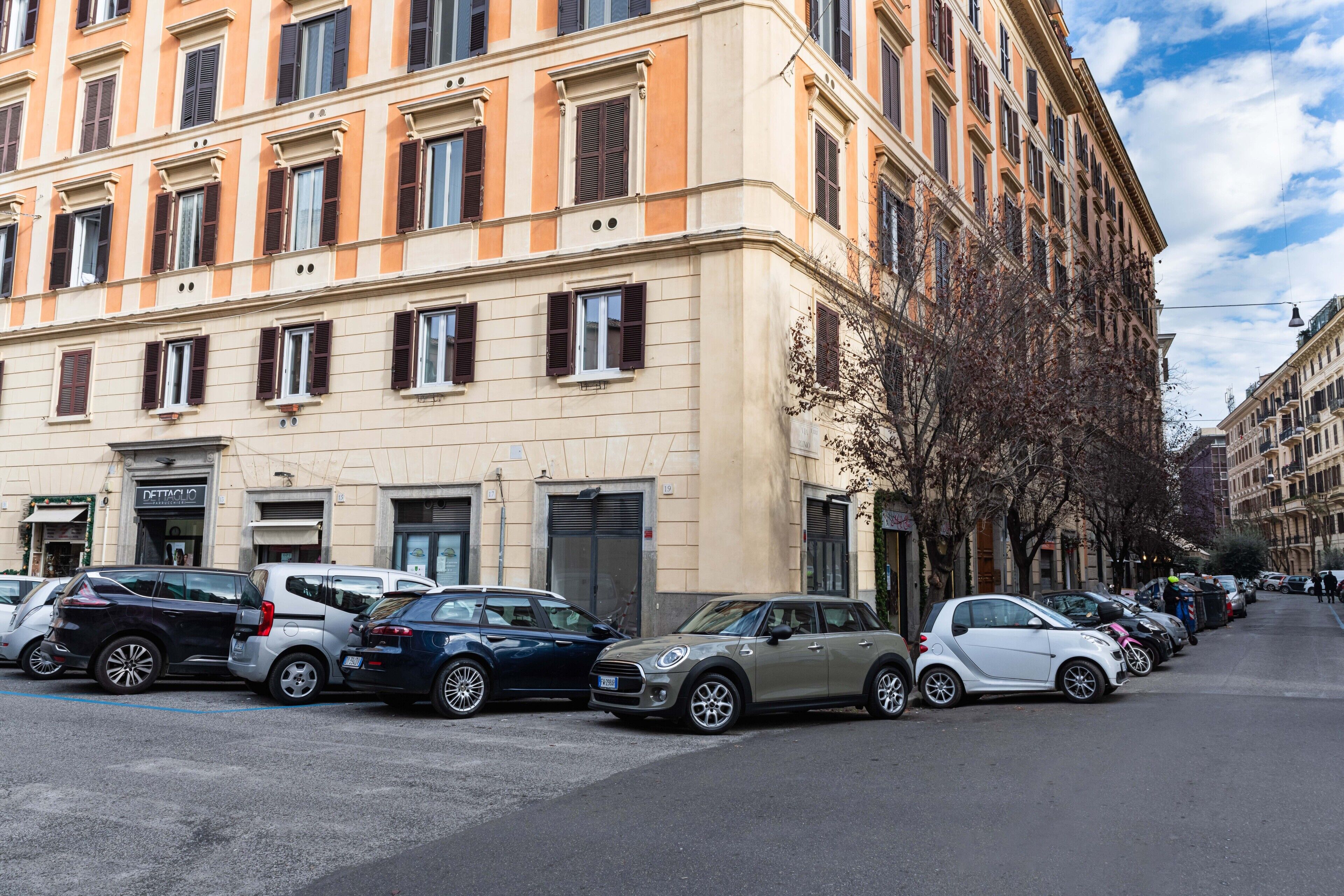 File di auto parcheggiate lungo una strada, con un edificio dotato di diverse finestre e un ingresso.