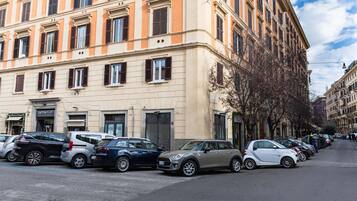 File di auto parcheggiate lungo una strada, con un edificio dotato di diverse finestre e un ingresso.
