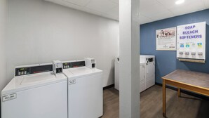 A laundry room with multiple washing machines, a table, and a wall poster advertising soap and bleach softeners. - HomeTowne Studios By Red Roof Newport News – Hampton West (Newport News)