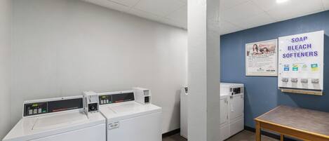 A laundry room with multiple washing machines, a table, and a wall poster advertising soap and bleach softeners.