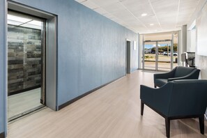 A modern hallway with a blue wall, wooden flooring, and two dark blue chairs. - HomeTowne Studios By Red Roof Newport News – Hampton West (Newport News)