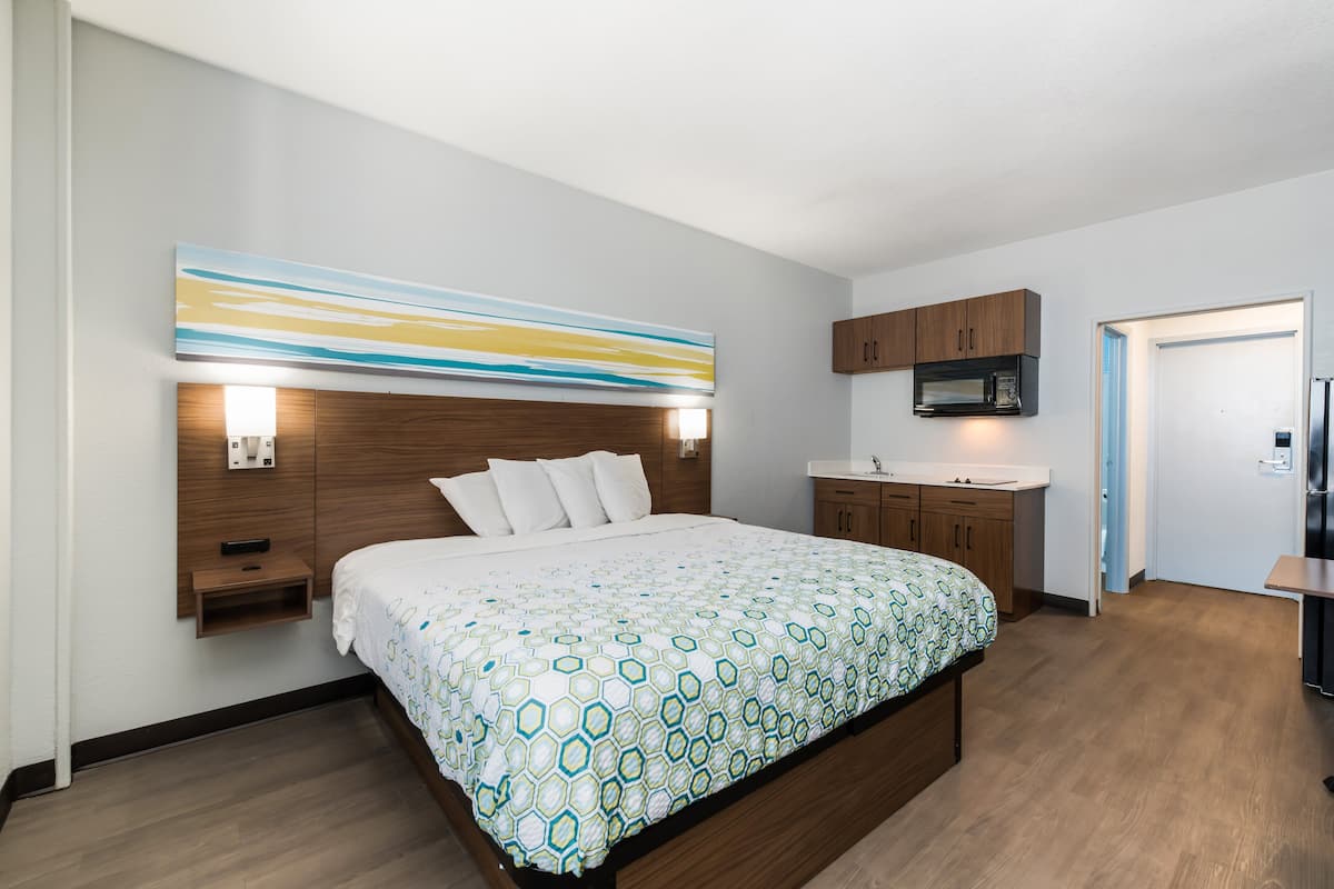A modern hotel room with a large bed, wooden headboard, a built-in TV, a microwave, and a small kitchenette.
