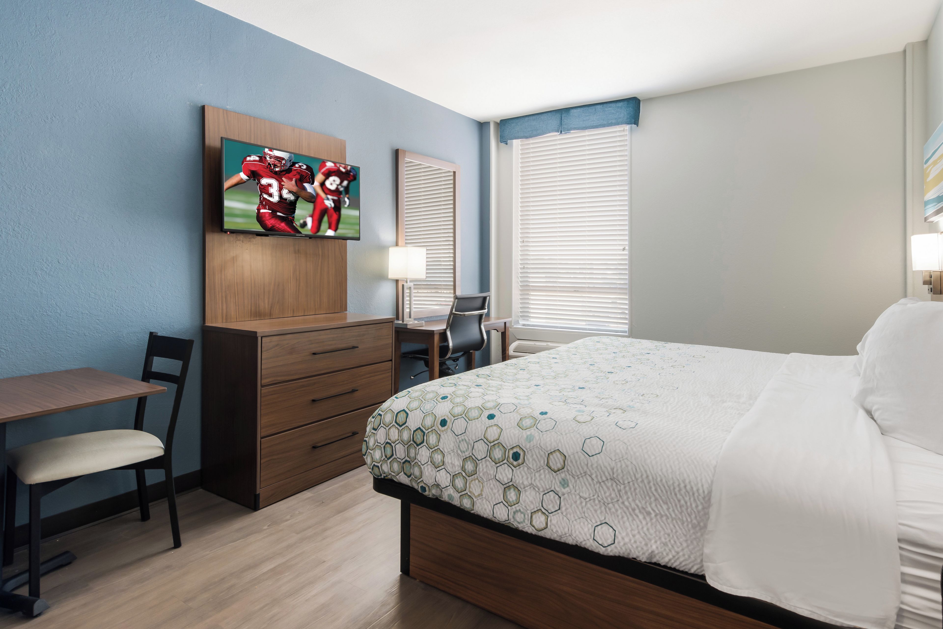 A hotel room with a bed, a desk, a chair, a TV mounted on the wall, and a window with blinds.