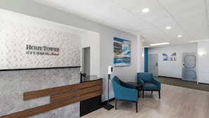 A reception area with a reception desk, two chairs, a wall-mounted artwork, and a door in the background. - HomeTowne Studios By Red Roof Newport News – Hampton West (Newport News)