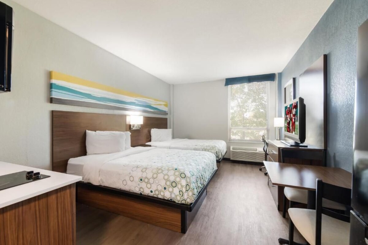 Habitación de hotel con dos camas, un escritorio, una silla, un televisor y una ventana con persianas.