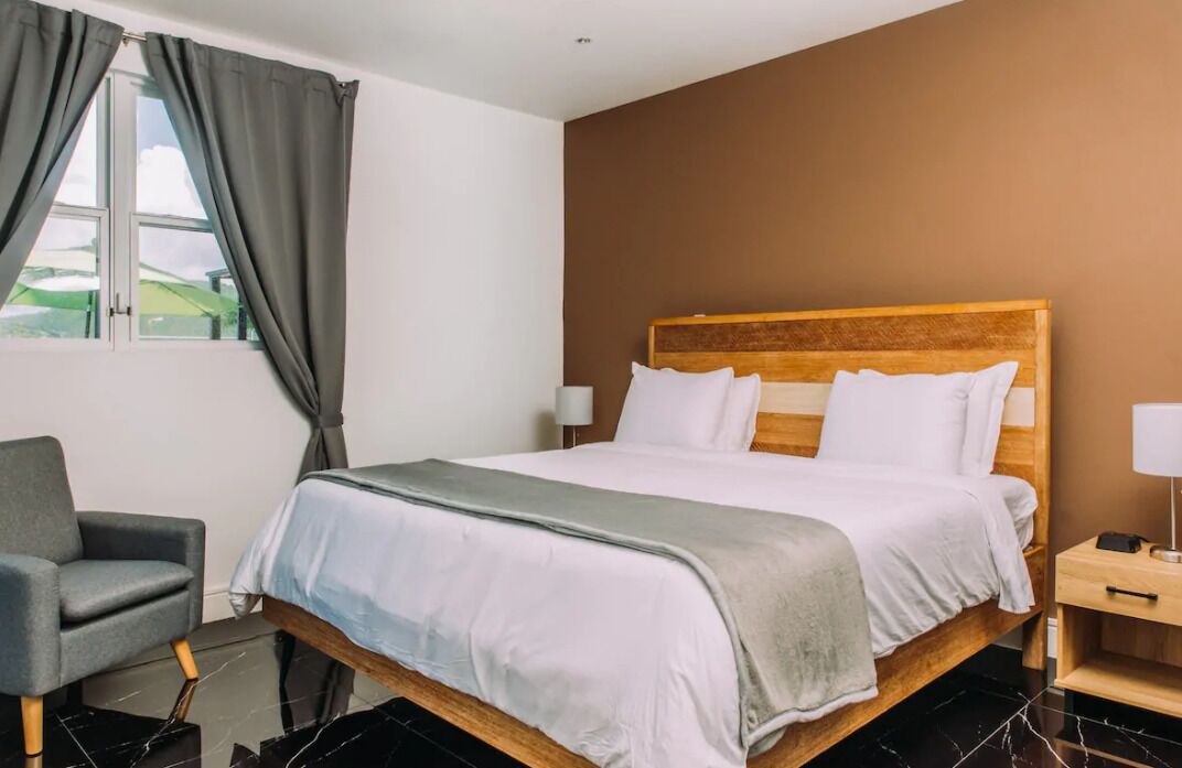 A hotel room with a wooden bed, a grey armchair, a bedside table with a lamp, and a window with curtains.
