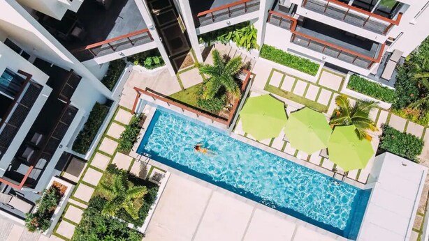 Ilmakuva modernista hotellista, jossa on suorakaiteen muotoinen uima-allas. Hotellia ympäröi vehreä kasvillisuus ja hyvin hoidettu puutarha.