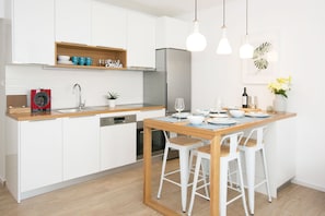 Una cocina moderna con armarios blancos, encimera de madera, horno empotrado, un fregadero y una zona de comedor con mesa de madera y taburetes blancos.