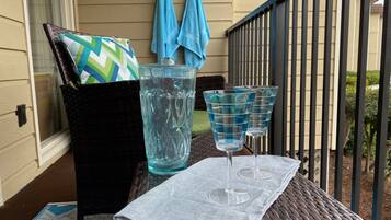 A wicker chair with a cushion, a glass pitcher, and two wine glasses on a table with a cloth.