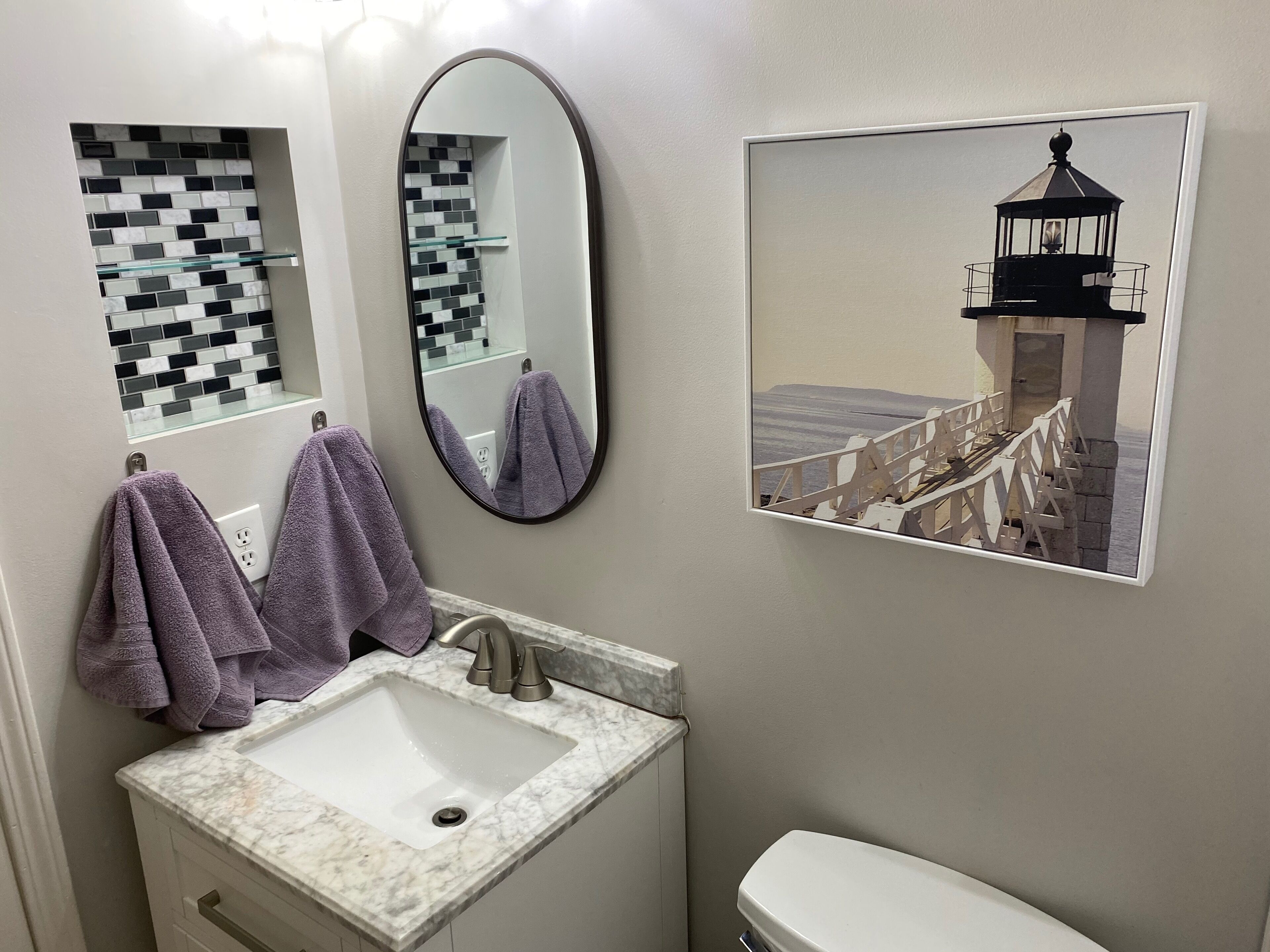 Un bagno con un lavandino bianco, uno specchio e un quadro appeso raffigurante un faro.