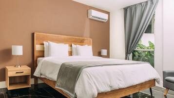 Una cama bien hecha con cabecera de madera, una mesita de noche con lámpara y vistas a zonas verdes a través de una ventana con cortinas.