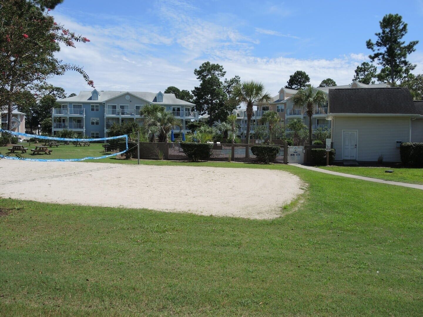 Ein Sandvolleyballfeld mit Picknicktischen, umgeben von einem Zaun und Palmen.
