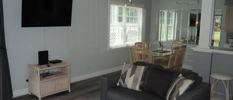Una sala de estar con un sofá gris, un televisor y un ventilador de techo. Hay una zona de comedor con una mesa y sillas, además de una cocina con mostrador y taburetes.