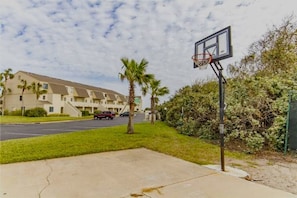 A basketball court with a hoop, surrounded by a parking lot and a building with multiple windows.