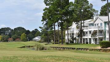 A large, multi-story building with balconies, situated on a golf course with a pond and trees.