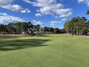 Un campo de golf con un estanque, árboles y casas al fondo.