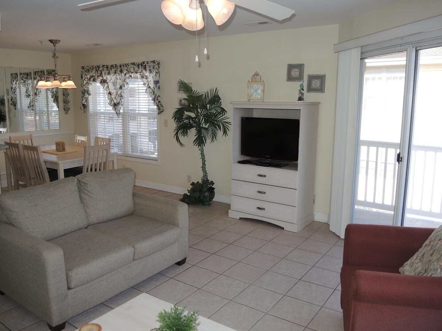 A living room with a grey sofa, a red sofa, a TV on a white stand, a dining area with a table and chairs, and a large window with blinds.