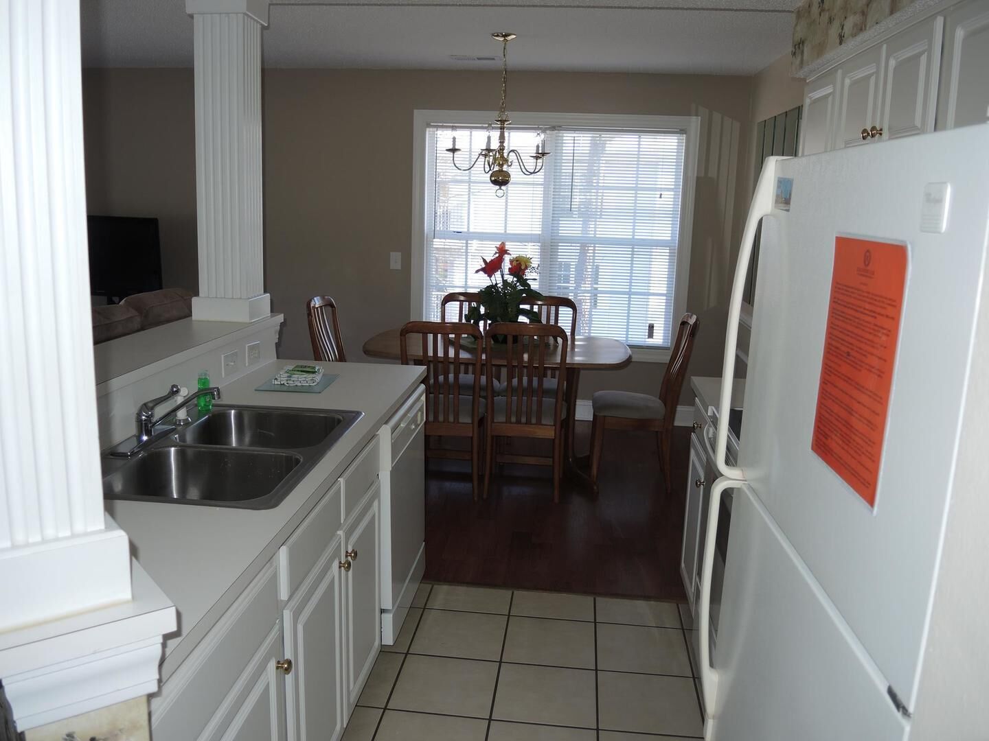 A compact kitchen with white cabinetry, a stainless steel sink, a dining table with chairs, a chandelier, and a refrigerator with a notice attached.