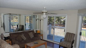 Un salon avec un canapé marron, un canapé beige, une table basse en bois et un fauteuil à motifs. On y trouve un ventilateur de plafond, une fenêtre avec des stores et une vue sur une terrasse extérieure.