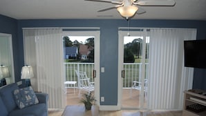 Un salon avec un canapé bleu, une table basse, un meuble pour la télévision et un ventilateur de plafond. Une porte-fenêtre coulissante donne accès à un balcon aménagé avec du mobilier d’extérieur.