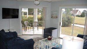 A living room with a blue sofa, a white coffee table, and a TV mounted on the wall. There is a sliding glass door leading to an outdoor patio area with chairs. - Family Friendly Golf Course View Condo 101M with Great Brunswick Plantation Resort Amenities by RedAwning (Calabash)