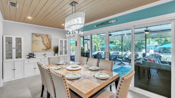 A dining area with a wooden table set for four, white cabinets, a map on the wall, and a view of the outdoor pool area. - Bayside Bliss 3 Bedroom Home by RedAwning (Anna Maria)