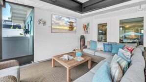 A modern living room with a large window, a wooden coffee table, a grey sofa with colorful cushions, and a painting of a beach scene. - Bayside Bliss 3 Bedroom Home by RedAwning (Anna Maria)