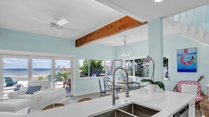 Una cocina moderna con una isla grande, un comedor con vista a la playa y una sala de estar con ventilador de techo y ventanas amplias.