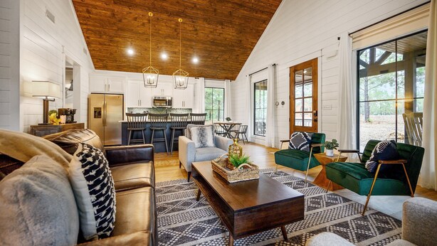 A spacious living room with a wooden ceiling, a kitchen in the background, and large windows.