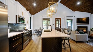 A modern kitchen with a central island, wooden ceiling, and a fireplace in the living area.