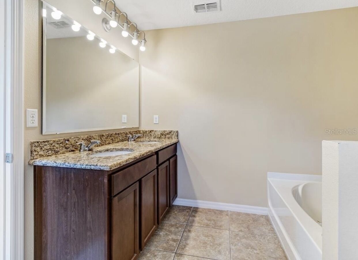 A bathroom with a double sink vanity, a large mirror with lights, and a bathtub.