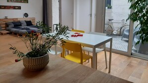 Un salon moderne avec un plancher en bois, une table à manger entourée de chaises jaunes, une plante en pot et un lit recouvert d’une couverture grise et bleue.
