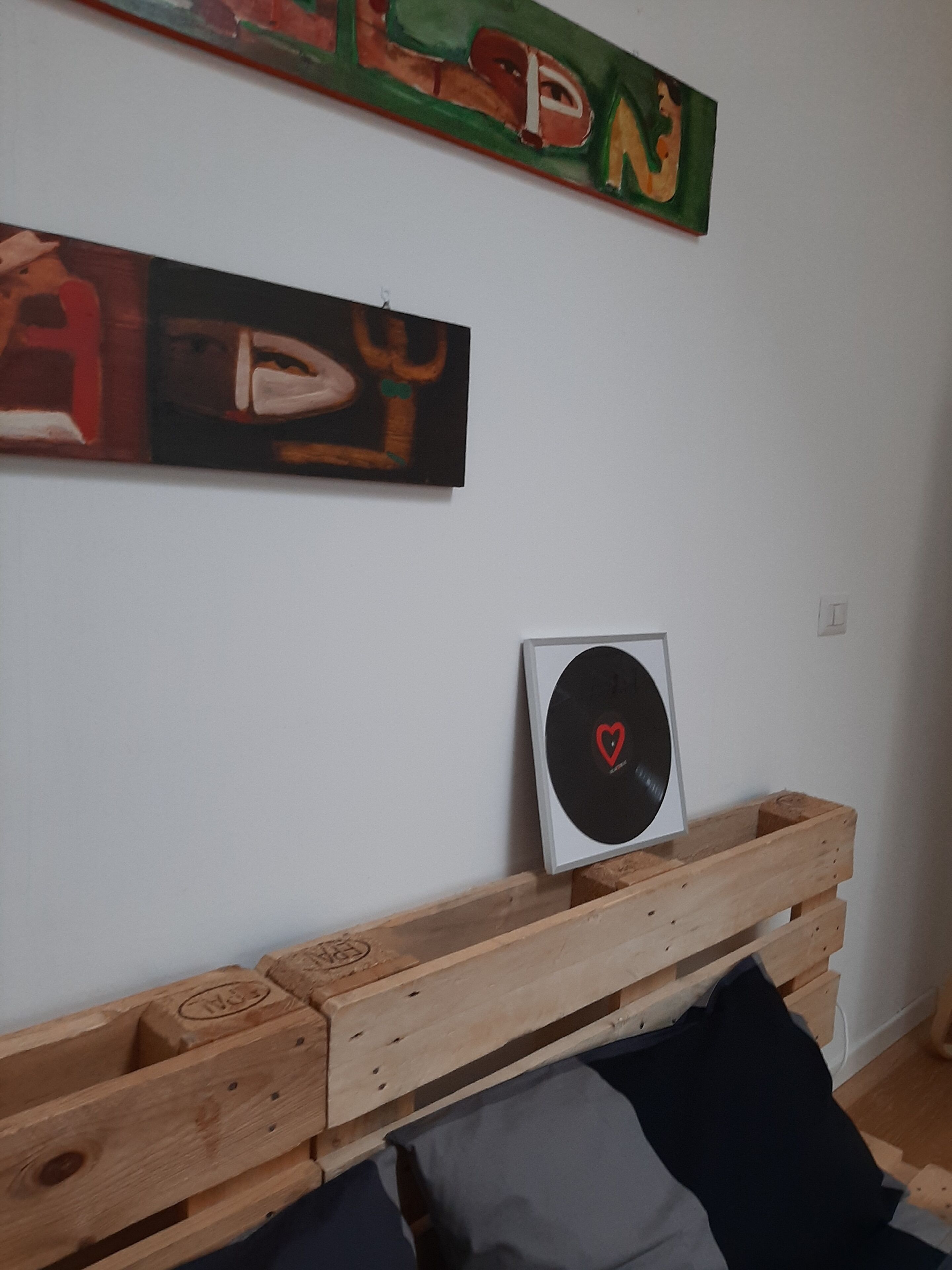 Une pièce avec un lit dont l’ossature est en bois, un tourne-disque encadré et deux tableaux abstraits accrochés au mur.