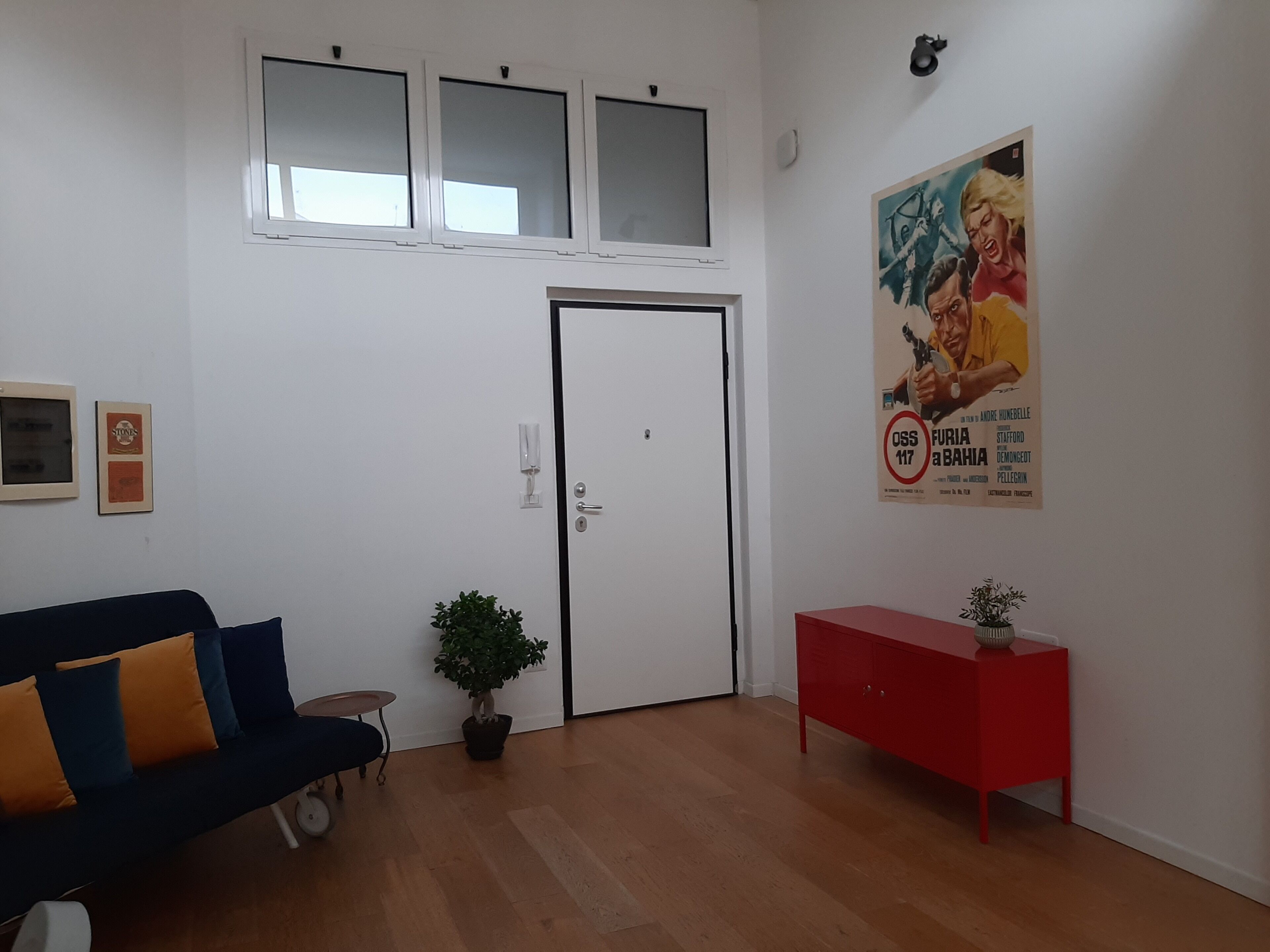 A room with a white door, a blue sofa with orange cushions, a small table, a potted plant, a red side table with a vase, and a poster on the wall.