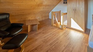 A room with a sloped ceiling, wooden flooring, a black leather chair, a small wooden table, and a wardrobe with a wooden staircase.