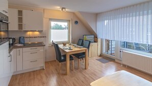 A compact kitchen with a dining area, wooden flooring, and a large window with curtains.
