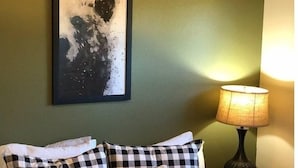 Ein Zimmer mit einem Bett, einem gerahmten abstrakten Gemälde, einer Lampe und karierten Kissen.