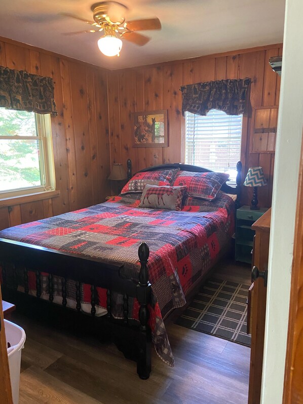 Une chambre à coucher aux murs lambrissés, avec un lit, un ventilateur de plafond, une fenêtre avec des stores et une table de chevet avec une lampe.