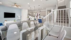 A modern living room with a glass dining table, grey upholstered chairs, a ceiling fan, recessed lighting, and a staircase with white railings.