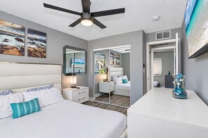 Une chambre à coucher comprenant un lit, des tables de chevet, un ventilateur de plafond, un miroir et des œuvres d’art accrochées aux murs.