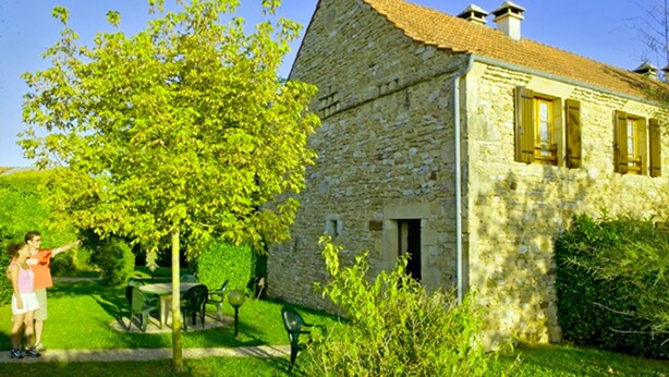 Una casa de piedra con techo de tejas, un pequeño jardín con una mesa y sillas, y un árbol.