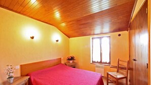 Un dormitorio con techo de madera, una cama con colcha roja, una mesita de noche con una planta, una silla y una ventana con persianas.