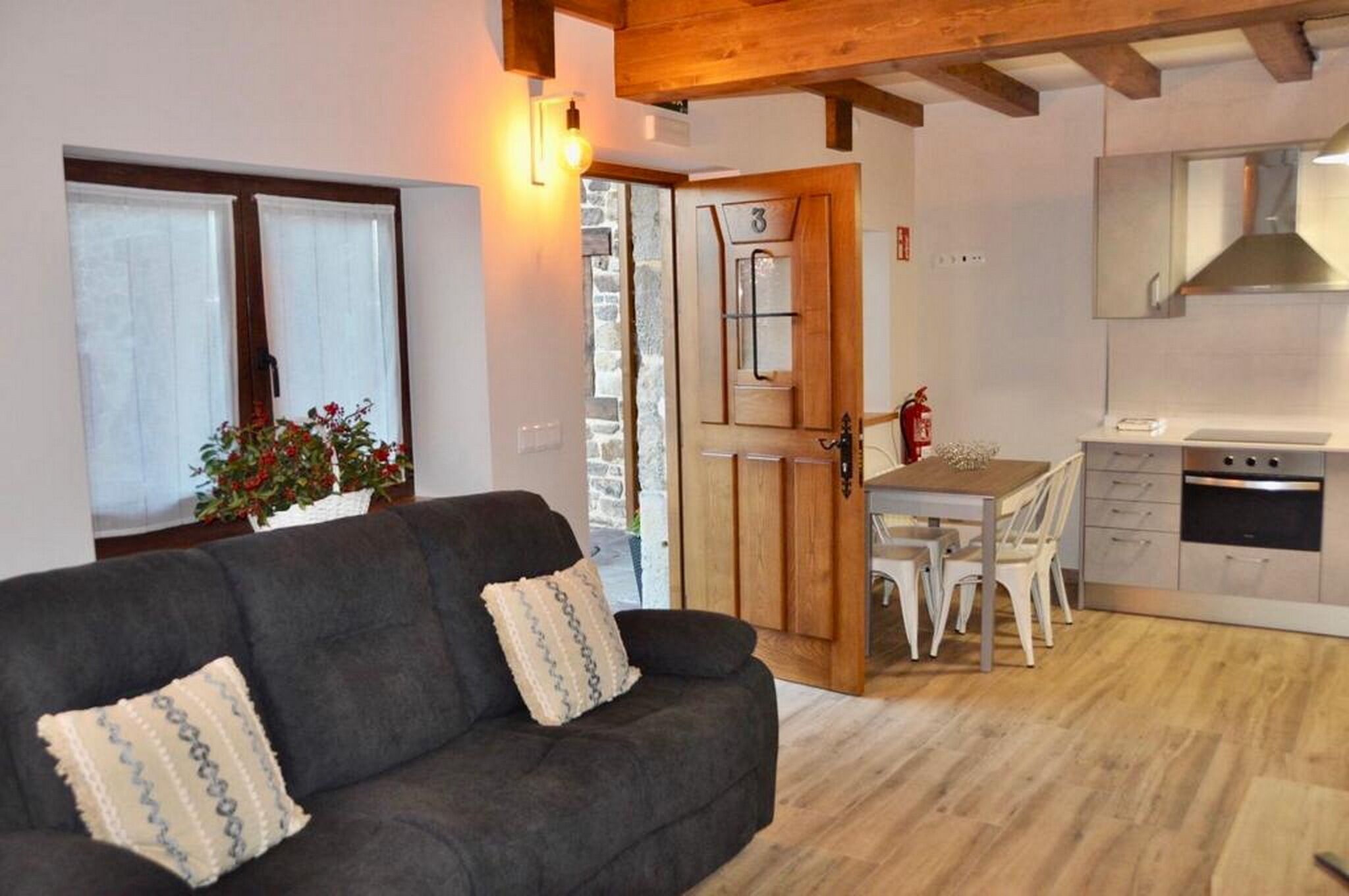 Una sala de estar acogedora con un sofá oscuro y una zona de comedor con mesa y sillas de madera. Se observa una cocina con electrodomésticos modernos al fondo.