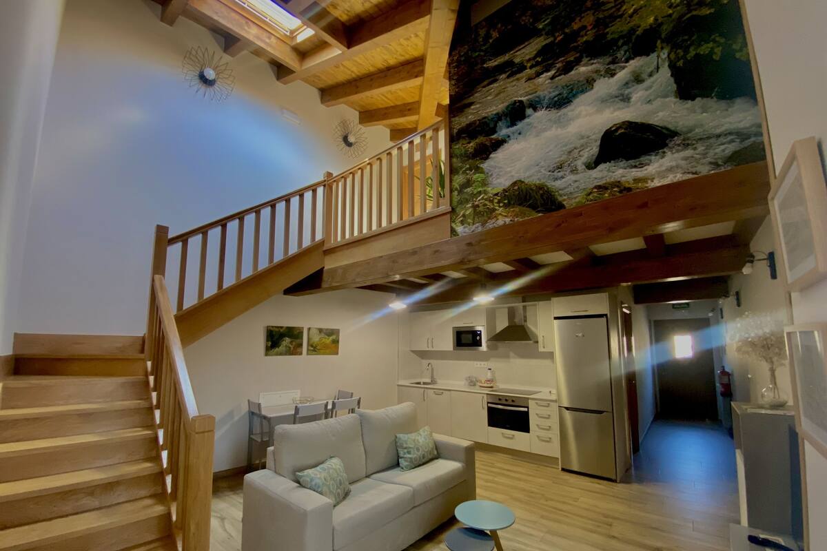 A spacious living area with a staircase leading to an upper level, a kitchen with modern appliances, and a large mural of a waterfall.