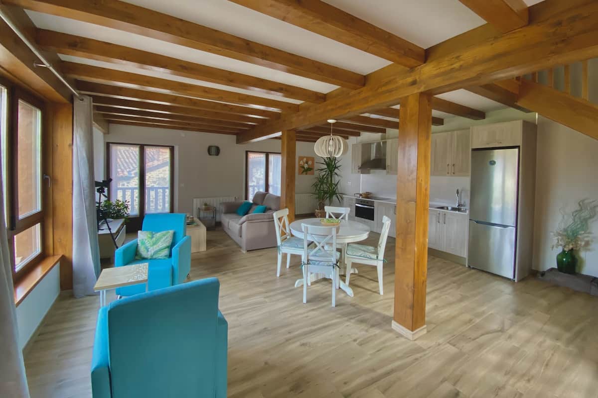 A spacious living area with wooden beams, a dining table with chairs, and a kitchen area with white cabinets and a refrigerator.